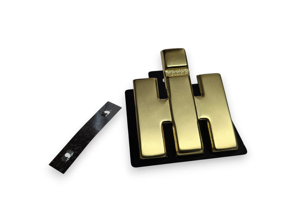 A gold-colored metal H-shaped badge or emblem with a black backing, shown alongside a separate black strip with two small silver dots, on a plain white background.